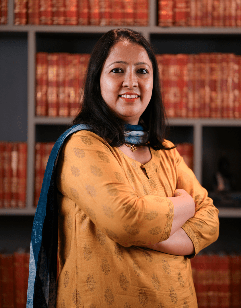A professional woman named Preksha Sharma wearing a suit, smiles confidently in front of a bookshelf filled with red and orange books.